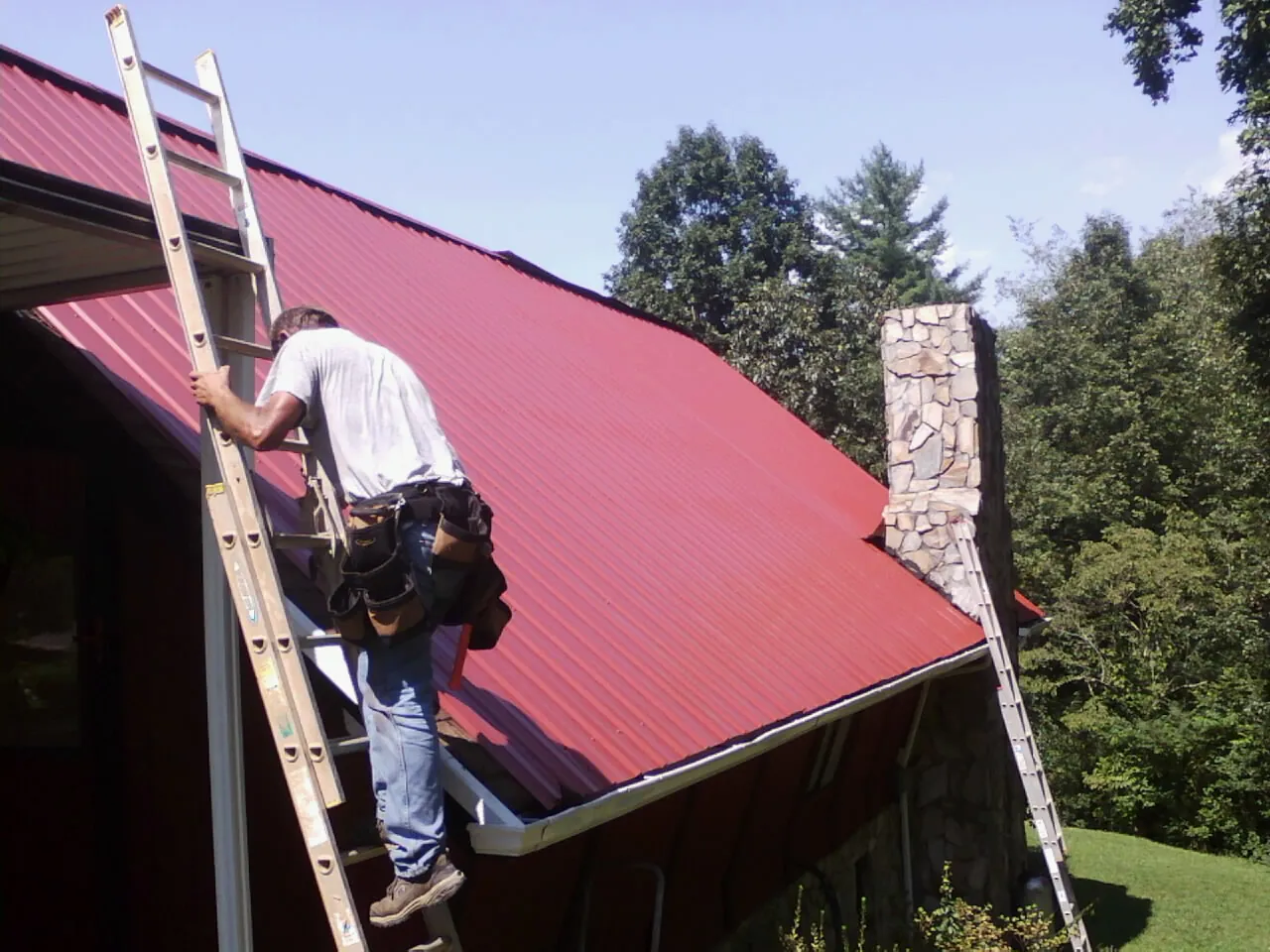 Roofer on a ladder beside a red metal roof and stone chimney — Soffit & Fascia Repair by Roofing Brothers in Colbert